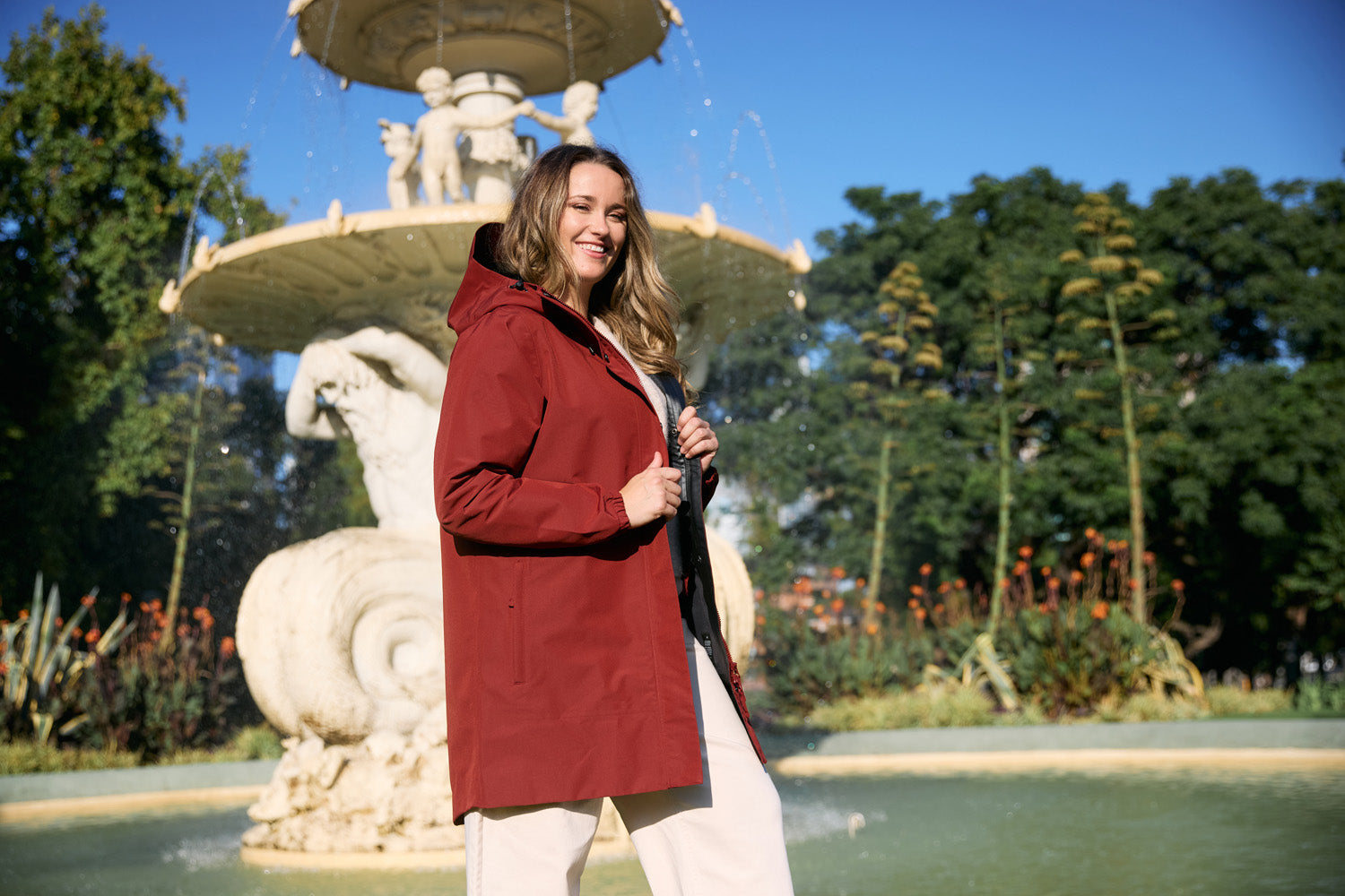 A model wearing the Refuge Jacket in Terracotta smiling happily in front of a fountain
