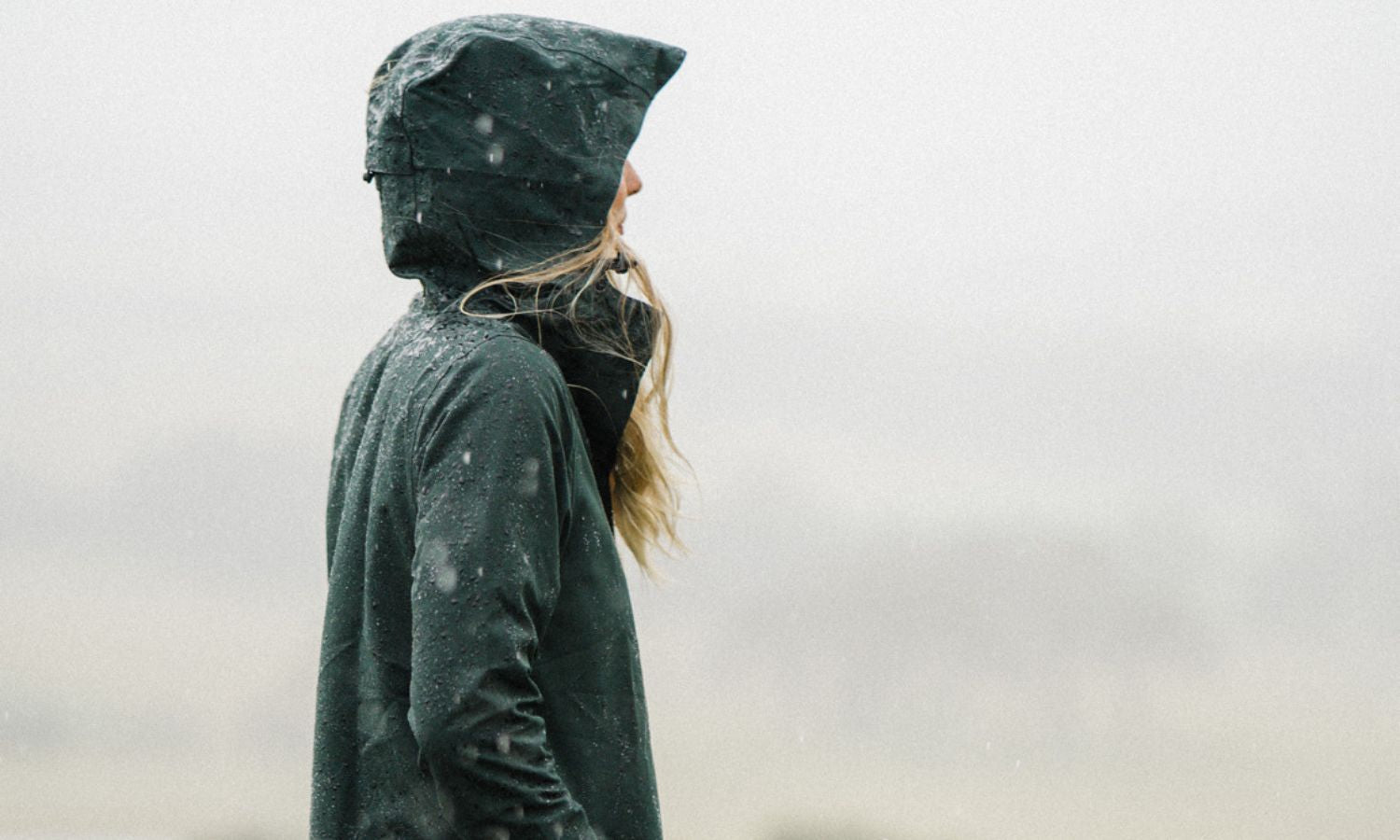 Woman in the rain, wearing the Elements jacket in Eucalyptus with the hood over her head