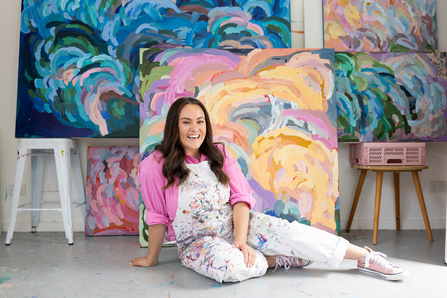 Erin Reinboth, the artist of the Fairy Bread Crumbs painting, sitting on the floor in front of her paintings wearing a colourful paint-stained apronsmiling happily